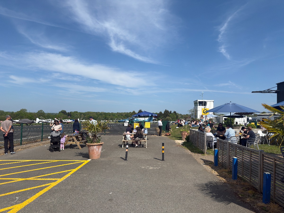 The Aerodrome Café terrace at Elstree EGTR on a blue-sky day, picnic benches and parasols, the white control tower with the yellow e logo, and light aircraft parked on the apron behind the fence.