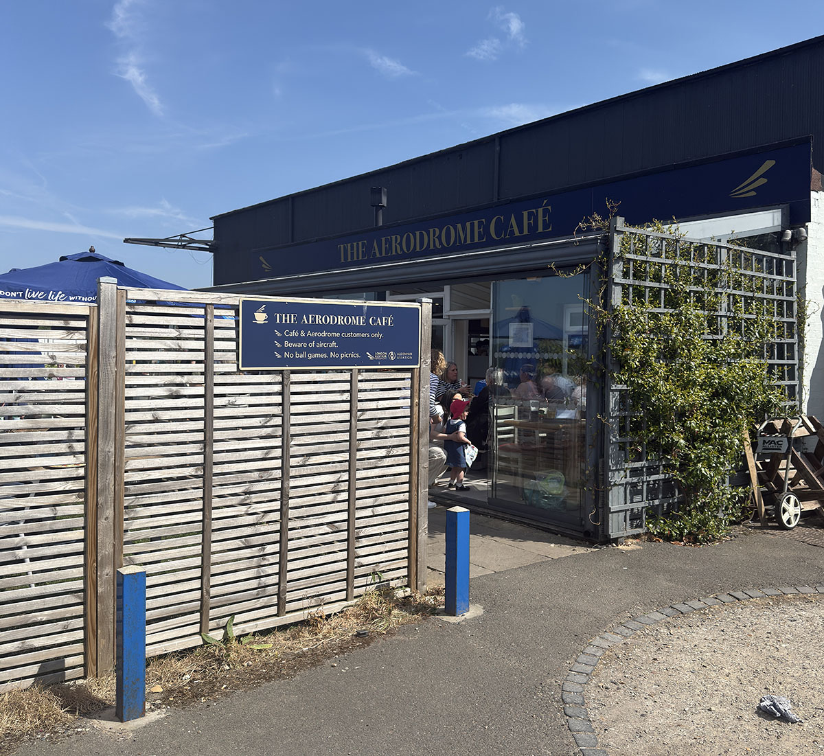 The Aerodrome Café frontage at Elstree EGTR, navy and gold signage with a house-rules board reading cafe and aerodrome customers only, beware of aircraft, no ball games, no picnics.