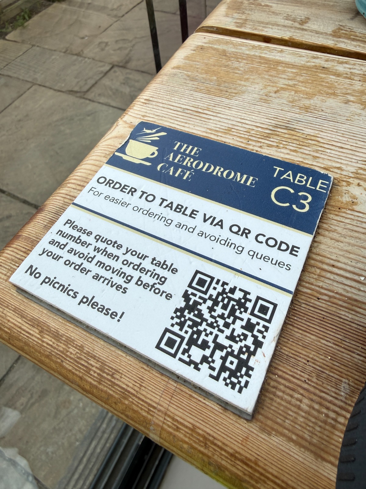 Order to table QR code card on a wooden bench at The Aerodrome Café, Elstree EGTR, marked Table C3 with the line no picnics please.