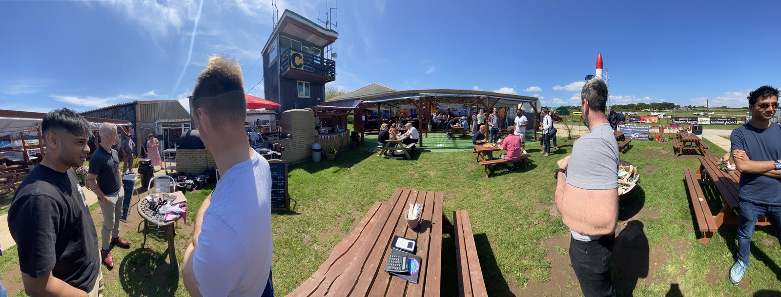 Busy outdoor seating at Sandown Airport bistro on a sunny day, with the control tower, covered seating and the Black Arrow rocket nose visible