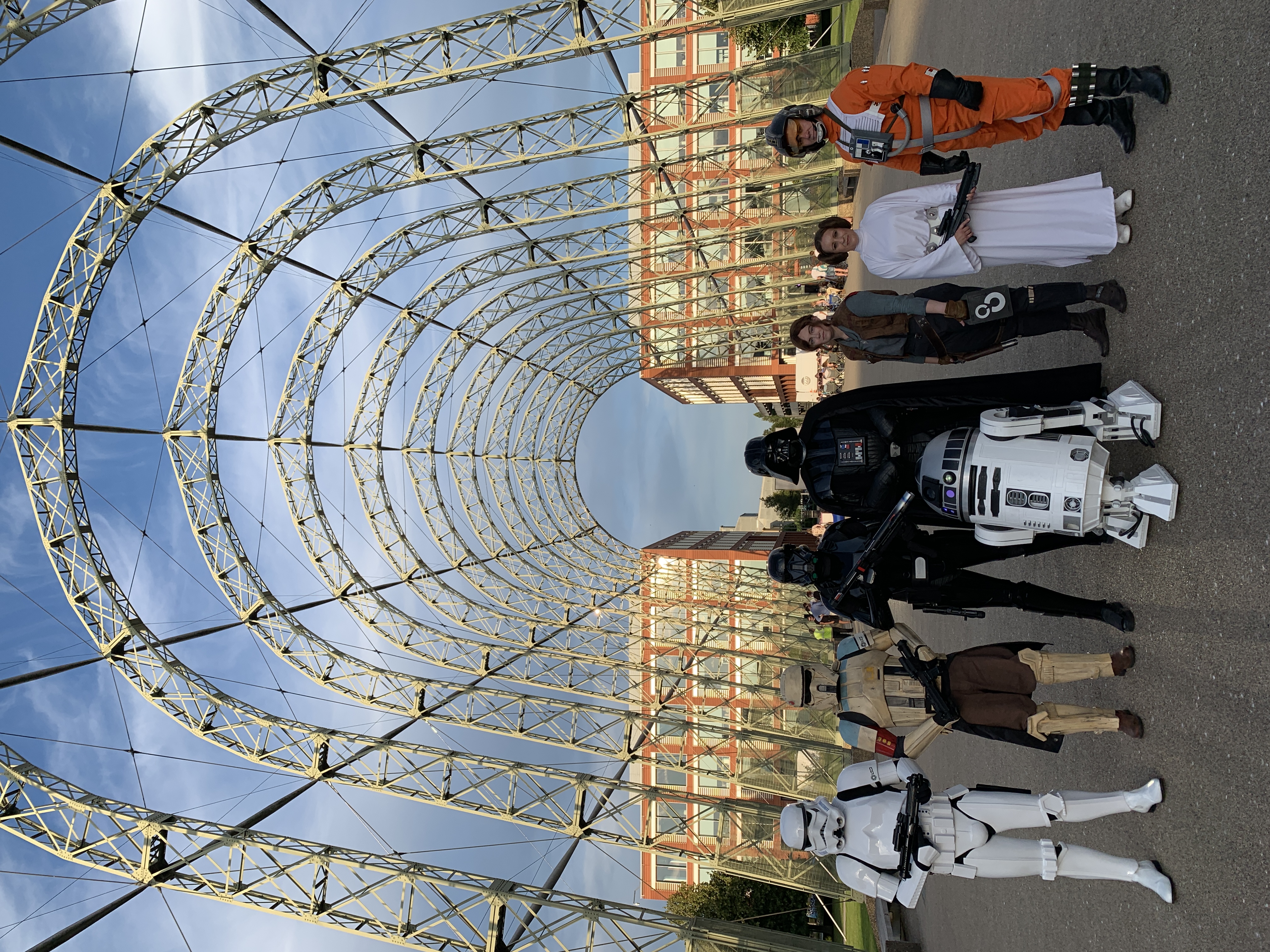 R2-TK and a Rogue One troop including Stormtrooper, Shoretrooper, Death Trooper, Darth Vader, Jyn Erso, Princess Leia and an X-Wing pilot, photographed under the exposed steel arches of Farnborough Airfield's historic hangar framework at a Rogue One screening, August 2019.